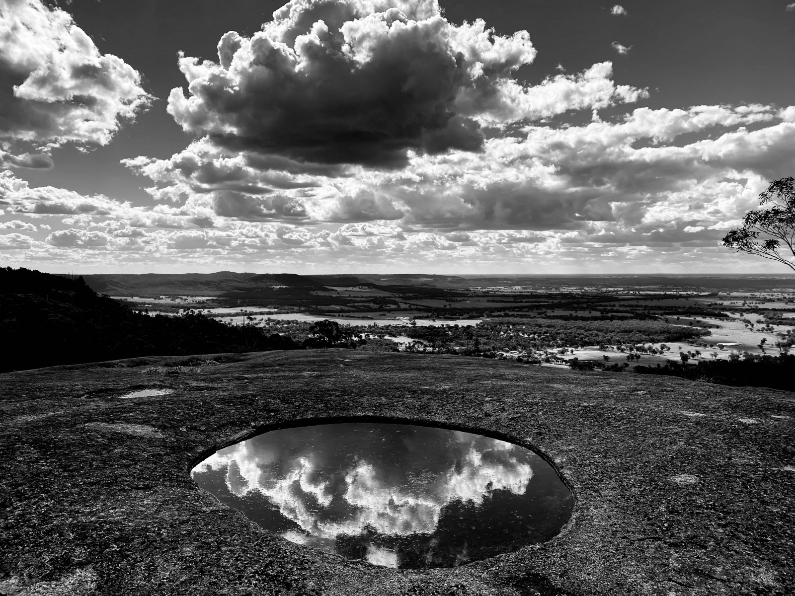 Black and white photo of a circular pool in a rocky surface, reflecting the clouds and sky above.